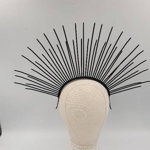May include: A black metal crown with a pointed design, resembling a sunburst. The crown is on a white mannequin head.