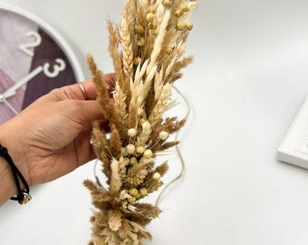 May include: A dried floral arrangement featuring a variety of beige, tan, and brown grasses and seed heads. The bouquet is tied with twine and held by a hand. The arrangement is likely for home decor or crafting.