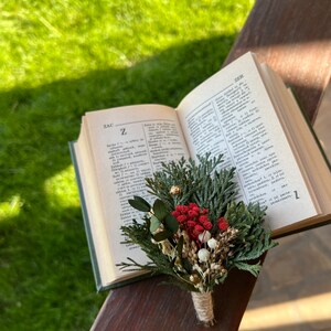 May include: A small, rustic boutonniere composed of dried greenery, red berries, and white accents, tied with twine. The boutonniere rests on an open book with the letter "Z" visible. The background features a wooden surface and green grass.