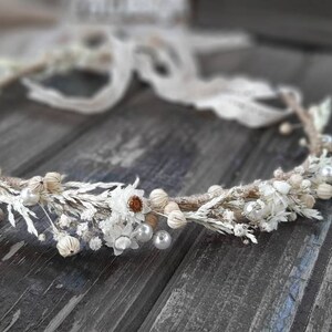 May include: A delicate floral crown featuring white dried flowers, small pearls, and seed pods. The crown is adorned with white daisies and is tied with a cream-colored lace ribbon. The rustic design is perfect for weddings or special occasions.