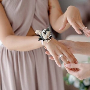 May include: A bridesmaid helps another bridesmaid put on a wrist corsage made of white flowers and green leaves. The corsage is tied with a white ribbon.