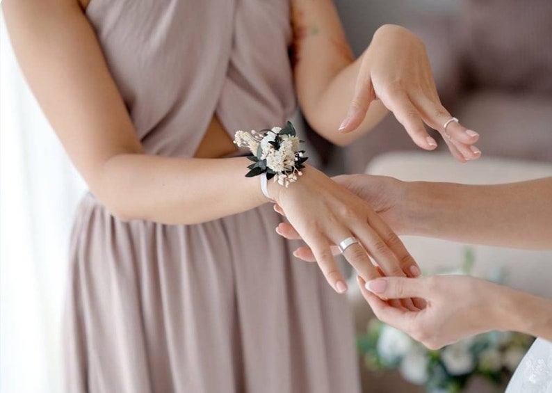 May include: A bridesmaid helps another bridesmaid put on a wrist corsage made of white flowers and green leaves. The corsage is tied with a white ribbon.