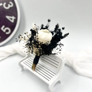 May include: A small bouquet of dried flowers with a white flower in the centre. The bouquet is tied with a black ribbon and is sitting on a white bench.
