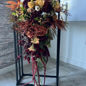 May include: A cascading bouquet of faux flowers in shades of burgundy, orange, and yellow. The arrangement includes roses, berries, and various foliage, displayed in a clear vase on a black metal stand. The bouquet is a mix of textures and colors.
