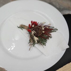 May include: A white napkin with a small bouquet of dried flowers and a pine cone. The bouquet includes red berries, brown pine cones, and green and brown foliage.