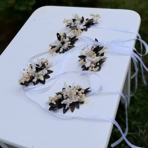May include: Five white and brown dried flower wrist corsages with white satin ribbon ties. The corsages are arranged on a white table.