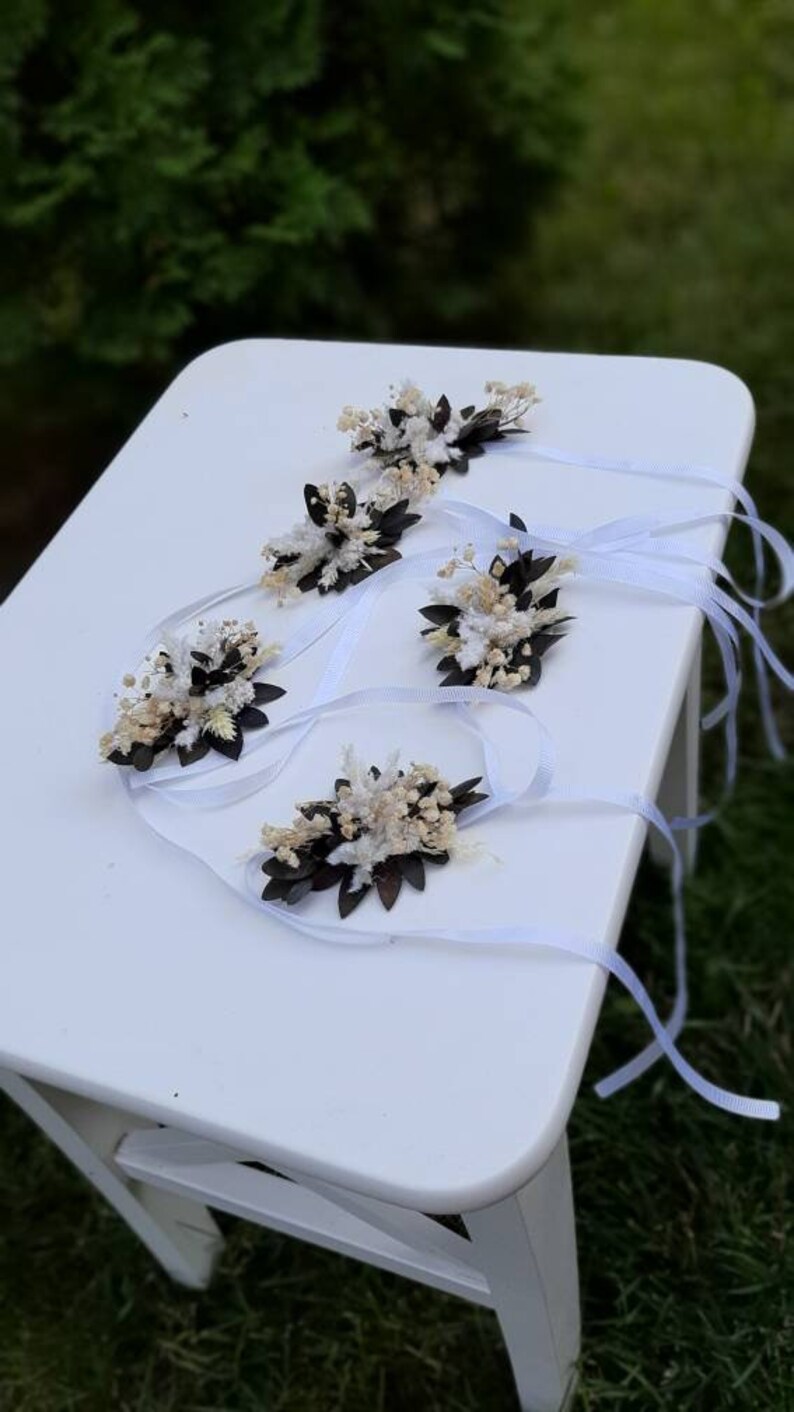 May include: Five white and brown dried flower wrist corsages with white satin ribbon ties. The corsages are arranged on a white table.