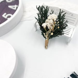 May include: A small bouquet of dried white flowers and green foliage tied with twine, sitting on a white bench with the text "Welcome to the World!" written on it.