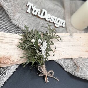 May include: A small bouquet of dried greenery and white flowers tied with twine. The bouquet is sitting on a piece of wood with a white sign that says "TriDesign" in the background.