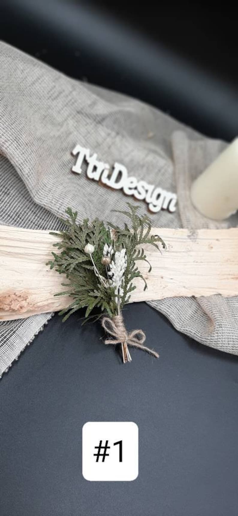 May include: A small bouquet of dried greenery and white flowers tied with twine. The bouquet is sitting on a piece of wood with a white sign that says "TriDesign" in the background.