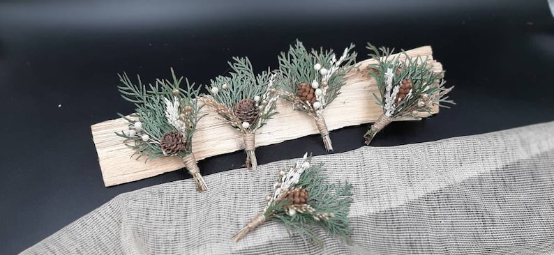 May include: Five small bouquets of greenery, pine cones, and white berries tied with twine. The bouquets are arranged on a wooden surface.
