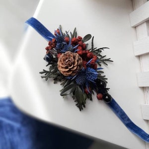 May include: A blue velvet ribbon with a dried floral arrangement featuring pine cones, blue thistle, and red berries.