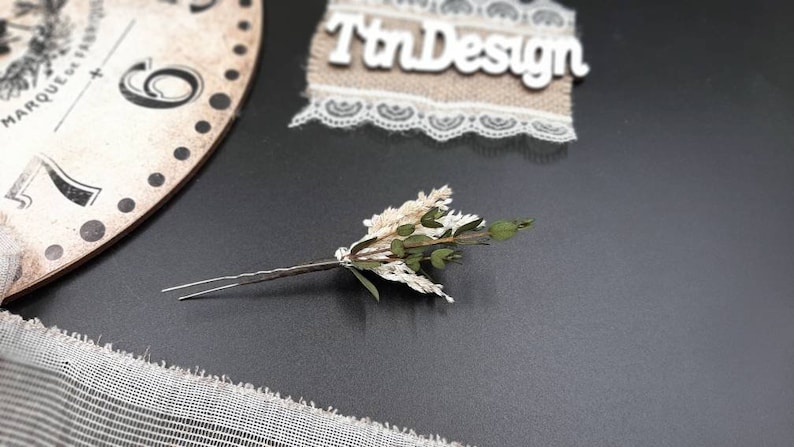 May include: A silver hair pin with a small bouquet of dried white and green flowers attached.