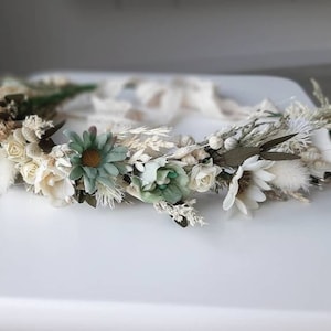 May include: A white and green floral crown with dried flowers and greenery. The crown is made with white roses, small white flowers, green leaves, and dried grasses.