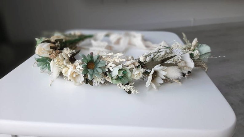 May include: A white and green floral crown with dried flowers and greenery. The crown is made with white roses, small white flowers, green leaves, and dried grasses.