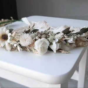 May include: A white and cream dried flower crown with a lace ribbon. The crown is made with various dried flowers, including daisies, roses, and grasses.