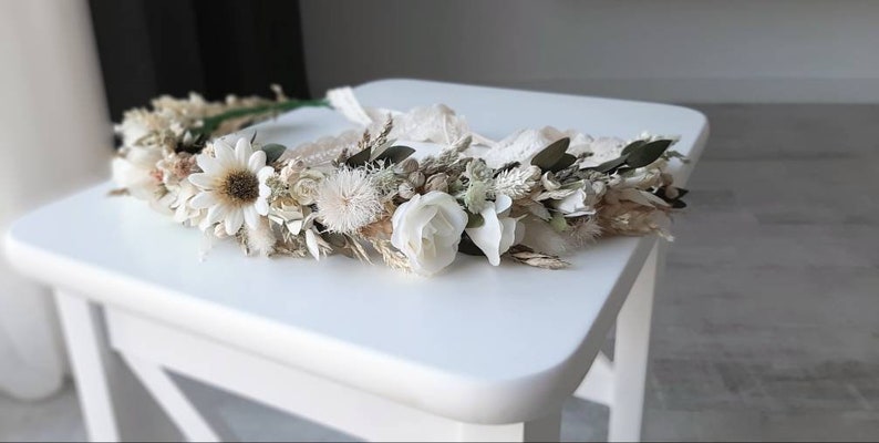 May include: A white and cream dried flower crown with a lace ribbon. The crown is made with various dried flowers, including daisies, roses, and grasses.