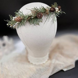 May include: A green and brown floral crown with pine needles, pine cones, and white berries. The crown is displayed on a white mannequin head.