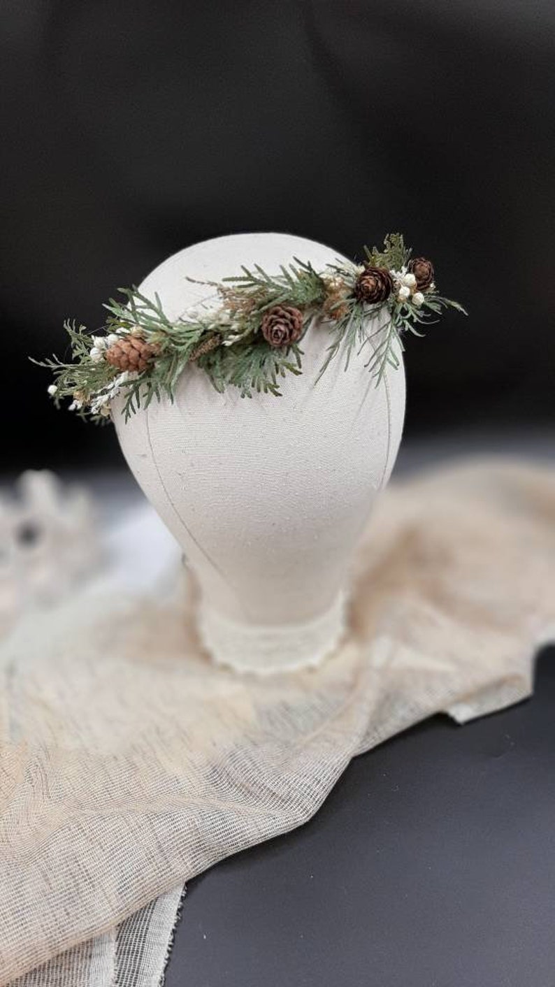 May include: A green and brown floral crown with pine needles, pine cones, and white berries. The crown is displayed on a white mannequin head.