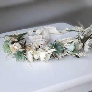 May include: A white and green floral crown with a lace ribbon. The crown is made of dried flowers and greenery.