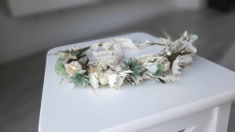 May include: A white and green floral crown with a lace ribbon. The crown is made of dried flowers and greenery.
