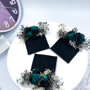 May include: Three black square cards are adorned with teal-colored roses and small white and brown dried flowers. The floral arrangements sit on a white circular surface, creating a striking contrast.