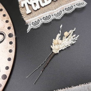 May include: A white dried flower hair pin with a brown wire stem. The pin is on a black surface.