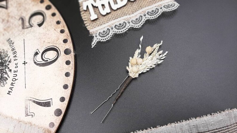May include: A white dried flower hair pin with a brown wire stem. The pin is on a black surface.