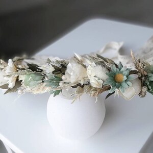 May include: A white and green floral crown with dried flowers and greenery. The crown is made with white and green flowers, dried grasses, and other natural elements.