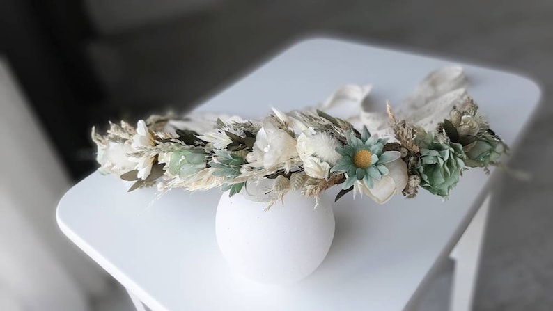 May include: A white and green floral crown with dried flowers and greenery. The crown is made with white and green flowers, dried grasses, and other natural elements.