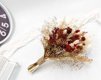 May include: A dried flower bouquet with a mix of beige, brown, and red dried flowers. The bouquet is tied with twine and is sitting on a white surface.