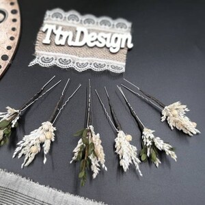 May include: Set of six hair pins with dried flowers. Each pin features a small cluster of dried white flowers and green leaves. The pins are arranged in a semi-circle on a black surface.