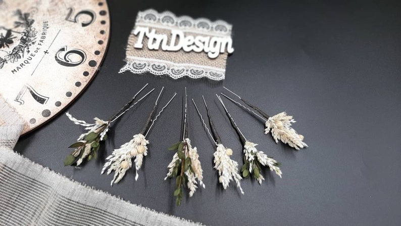 May include: Set of six hair pins with dried flowers. Each pin features a small cluster of dried white flowers and green leaves. The pins are arranged in a semi-circle on a black surface.
