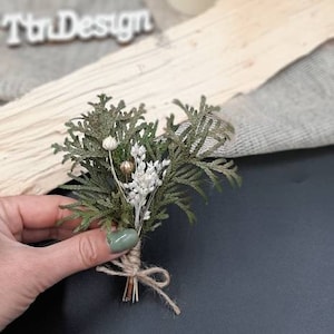 May include: A small bouquet of dried greenery and white flowers tied with twine. The bouquet is held in a hand with green nail polish.