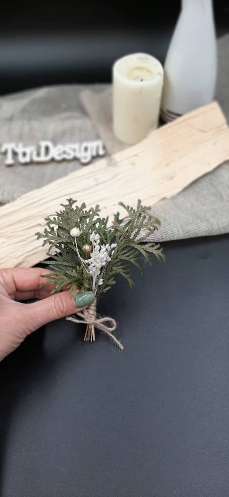May include: A small bouquet of dried greenery and white flowers tied with twine. The bouquet is held in a hand with green nail polish.