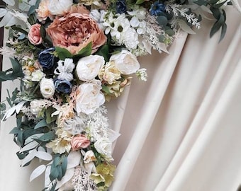 Op de afbeelding: Een waterval van bloemen met een grote, roestkleurige pioenroos als middelpunt, omringd door witte, roze en marineblauwe bloemen en weelderig groen blad. De opstelling staat tegen een crèmekleurige achtergrond.