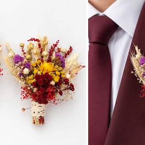 May include: A boutonniere featuring a cluster of dried flowers including yellow, purple, and deep red blooms, accented with stalks of wheat and small seed pods. The stems are wrapped in a cream-colored lace ribbon and secured with a pin.