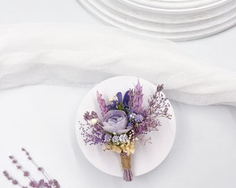 Op de afbeelding: Een klein boeket gedroogde bloemen in tinten van paars en wit, gebonden met touw, op een wit rond bord. Het boeket bestaat uit lavendel, gipskruid en andere kleine bloemen. De tekst "TTNdesign" staat op een houten bord onder het boeket.