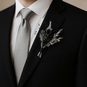 Könnte beinhalten: Schwarzer Anzug, silberne Krawatte und weißes Hemd. Eine kleine, dunkle Blumen-Boutonniere ist am Revers befestigt. Das Bild ist eine Nahaufnahme des Oberkörpers, die sich auf die formelle Kleidung konzentriert.