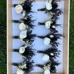 Pode incluir: Dez boutonnières de flores secas brancas e pretas dispostas em uma caixa. Cada boutonnière apresenta uma flor branca, ervas secas pretas e penas pretas.