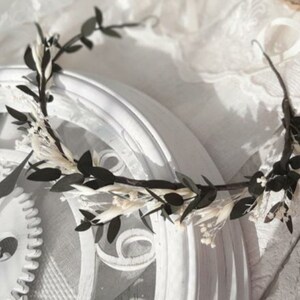 May include: A black and white floral crown with small white flowers and dark green leaves. The crown is resting on a white surface.