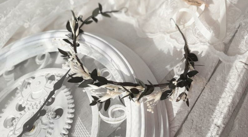 May include: A black and white floral crown with small white flowers and dark green leaves. The crown is resting on a white surface.