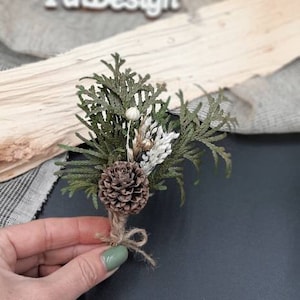 May include: A small bouquet of dried greenery, including cedar, pine cones, and white flowers, tied with twine. The bouquet is held in a hand against a dark background.