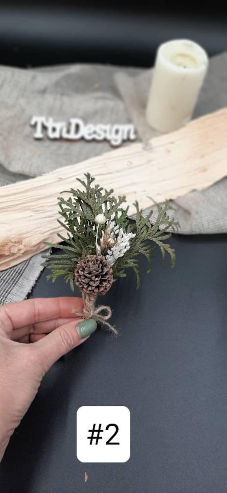 May include: A small bouquet of dried greenery, including cedar, pine cones, and white flowers, tied with twine. The bouquet is held in a hand against a dark background.