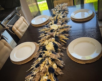 Op de afbeelding: Een tafelsetting met een lang, gedroogd bloemstuk in beige, wit en blauwe tinten. Het bloemstuk is geplaatst op een donkerhouten tafel met witte borden en geweven placemats.