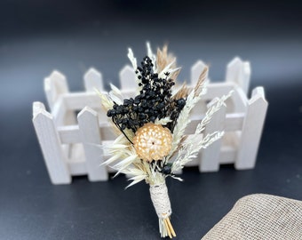 May include: A small bouquet of dried flowers with black berries, white pampas grass, and a beige dried flower. The bouquet is tied with a white lace ribbon and is sitting on a white picket fence.