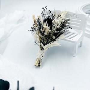 Könnte beinhalten: Ein kleiner Strauß getrockneter Blumen in Weiß- und Schwarztönen, gebunden mit einem weißen Spitzenband. Der Strauß liegt auf einer weißen Holzbank.
