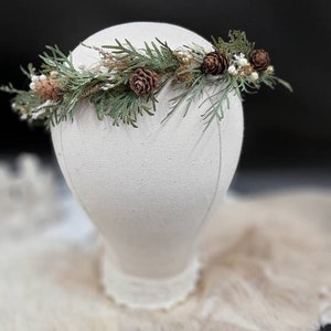 May include: A green and brown floral crown with pine cones and small white flowers. The crown is on a white mannequin head.