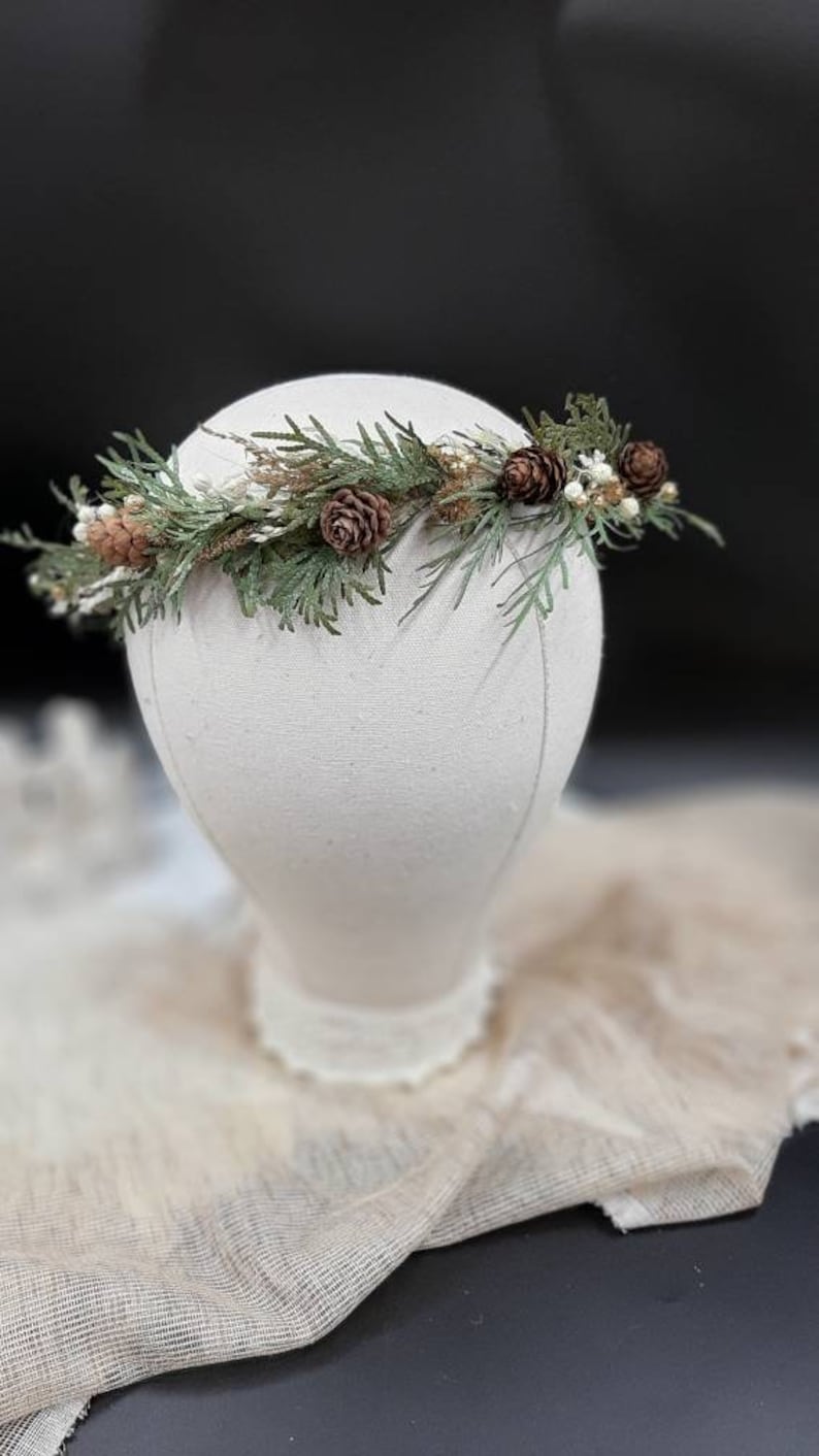 May include: A green and brown floral crown with pine cones and small white flowers. The crown is on a white mannequin head.
