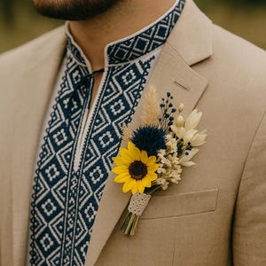 Op de afbeelding: Een beige blazer met een wit en marineblauw geborduurde overhemdkraag. Een gele zonnebloem en andere gedroogde bloemen zijn op de revers van de blazer gespeld. Het overhemd heeft een geometrisch patroon.